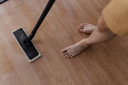 High-angle view of barefoot male cleaning hardwood floor with steam mop during house cleaning, close-up. Maintaining hygiene and cleanliness in home environment. Concept of cleaning and household.の写真素材