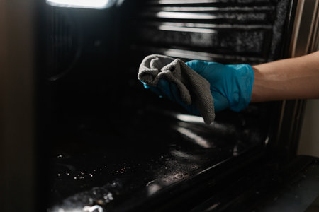 Professional cleaner worker wearing blue rubber gloves wiping inside of oven with gray rag, ensuring hygiene, cleanliness in kitchen, close-up. Concept of domestic work, housekeeping and household.の写真素材