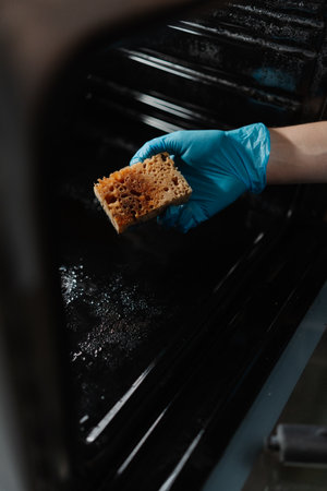 Vertical cropped shot of male hand in blue rubber glove holding dirty sponge, scrubbing soiled gas oven covered in grease stains and splashes, tackling mess in kitchen environment, close-up.の写真素材