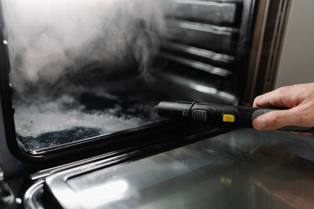 Professional cleaner male using steam cleaner to remove grease and grime from inside surface of modern gas oven, ensuring spotless and hygienic kitchen appliance. Concept of hygiene and cleanliness.の写真素材
