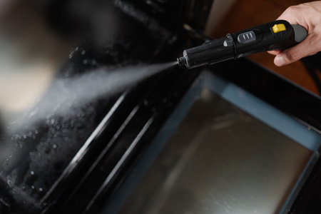 Top view of professional cleaner male sanitizing black gas oven interior with high-temperature steam, demonstrating kitchen cleaning hygiene process, close-up. Concept of cleanliness and housekeeping.の写真素材