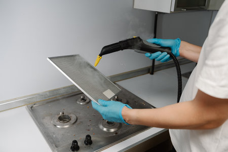 Housewife male wearing protective gloves sanitizing kitchen extractor filter using high-pressure steam cleaning equipment over gas stove. Concept of hygiene and cleanliness in domestic environment.の写真素材