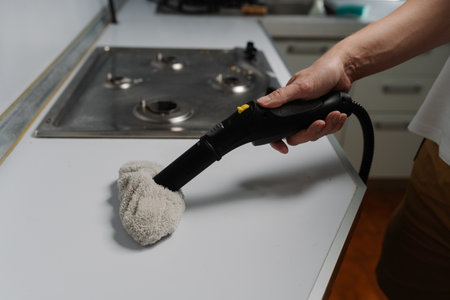 Close-up cropped shot of unrecognizable man wiping kitchen countertop with sanitizing steam cleaner near stovetop, maintaining spotless domestic hygiene. Concept of maintenance modern home appliance.の写真素材