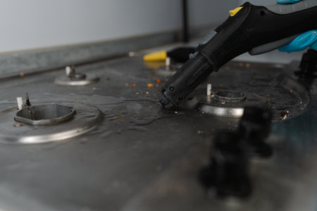 Cleaning professional wearing blue gloves using steam cleaner to remove burned-on food and grease from dirty gas stove, ensuring hygiene and cleanliness in kitchen setting, close-up.の写真素材