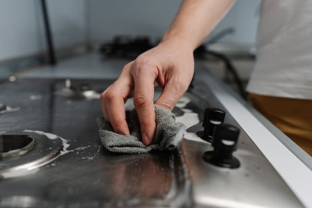 Housewife male using rag and detergent cleaning stainless steel gas stove, ensuring hygiene and maintenance in kitchen, close-up cropped shot. Concept of domestic work, housekeeping and household.の写真素材