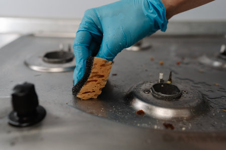 Close-up of cleaning service worker in gloves scrubbing dirty gas stove with sponge, removing grease and food remnants ensuring clean and hygienic kitchen. Concept of household chores and hygiene.の写真素材