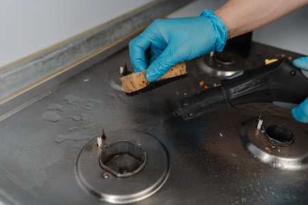 Homemaker male wearing blue glove cleaning dirty gas stove, removing grease and food remnants with sponge and detergent, close-up. Concept of household chores and kitchen hygiene.の写真素材