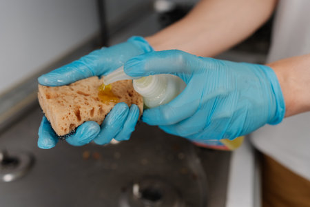 Gloved hands squeezing yellow dish soap onto sponge, getting ready to cleaning kitchen gas stove, ensuring hygienic and spotless environment. Concept of domestic work, housekeeping and household.の写真素材