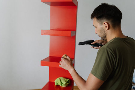 Back view of young man using steam cleaner and green microfiber cloth sanitizing red wooden shelf, performing housekeeping service for spotless and hygienic home environment.の写真素材