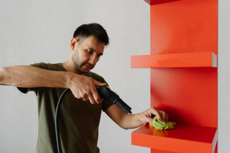 Portrait of focused young man cleaning red shelf using steam cleaner and sponge, ensuring hygiene and spotless surface in domestic setting. Concept of domestic work, housekeeping and household.の写真素材