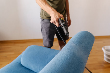 Cleaning technician sanitizing blue sofa with high-temperature steam cleaner in contemporary living...