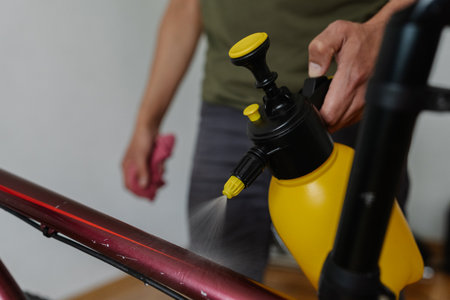 Close-up cropped shot of unrecognizable man cleaning bicycle frame, spraying water from yellow-black bottle, holding cloth in hand, preparing professional bike maintenance. Concept of bike service.の写真素材