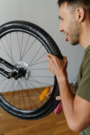 Funny cyclist male jokingly licking tire of mountain bike wheel while holding rag, showcasing humorous approach to bicycle maintenance, vertical portrait. Concept of bicycle transport service.の写真素材
