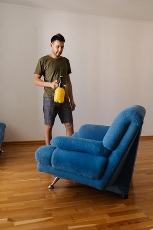 Full length vertical portrait of young man spraying water using yellow plastic sprayer on blue sofa in stylish living room, engaging home cleaning and maintenance. Concept of household cleanliness.の写真素材