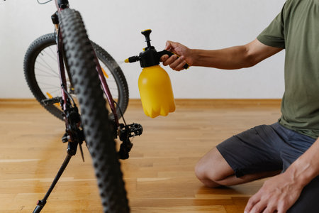 Side view of cyclist cleaning wheel of mountain bike using yellow water sprayer bottle, ensuring proper maintenance and hygiene for optimal performance at home. Concept of bike service, close-up.の写真素材