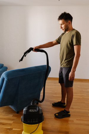 Side view of handsome bearded young man in casual clothes removing dirt and stains using washing vacuum cleaner to clean blue sofa in living room, demonstrating modern approach to household chores.の写真素材