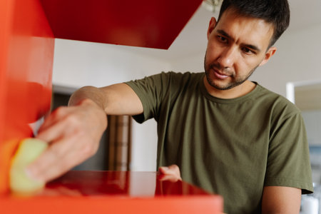 Portrait of focused janitor using cleaning cloth, wiping dusty dirty red shelf showcasing commitment to cleanliness and hygiene, close-up. Concept of domestic work, housekeeping, household.の写真素材