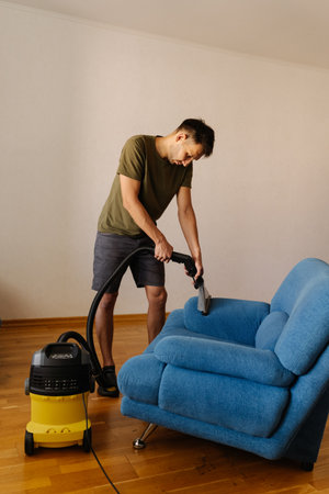 Full length vertical portrait of man in causal clothing using washing vacuum cleaner to clean blue sofa in home setting, demonstrating modern approach to household chores and upholstery maintenance.の写真素材