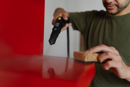 Housewife male using modern appliance to housekeeping, cleaning red shelf using steam cleaner and sponge, ensuring hygiene and spotless surface, close-up. Concept of hygiene and cleanliness.の写真素材