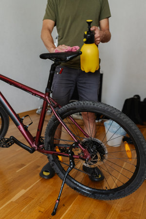 Vertical shot of cyclist male cleaning saddle of mountain bike using spray bottle and cloth, ensuring spotless and ready for next ride at home, close-up. Concept of bicycle maintenance and service.の写真素材