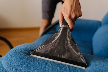 Detail cropped shot of unrecognizable cleaning service worker male using wet vacuum cleaner to remove dirt and stains from blue sofa, ensuring hygienic cleaning process. Concept of housekeeping.の写真素材