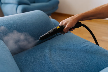 Cleaning service worker holding steam cleaner machine and cleaning blue sofa, removing dirt and dust with hot steam in living room, close-up. Concept of hygiene and cleanliness in domestic environmentの写真素材