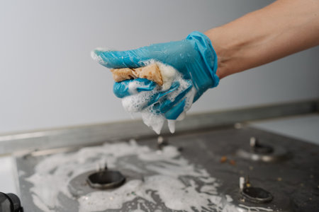 Cleaning service employee wearing protective gloves squeezing soapy water from sponge during cleaning dirty gas stove, to removing stubborn grime and grease. Concept of domestic work and household.の写真素材