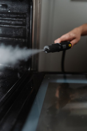 Vertical cropped shot of cleaner person sanitizing black gas oven interior with high-temperature steam, demonstrating kitchen cleaning hygiene process, close-up. Concept of cleanliness, housekeeping.の写真素材