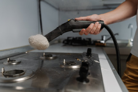 Closeup of housekeeper cleaning dirty gas stove using steam cleaner and cloth, removing grease and burnt food residues, ensuring hygienic kitchen environment. Concept of modern housekeeping appliance.の写真素材