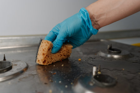 Housewife wearing rubber gloves scrubbing dirty gas stove with sponge, removing grease and food remnants ensuring clean and hygienic kitchen. Concept of household chores and hygiene.の写真素材