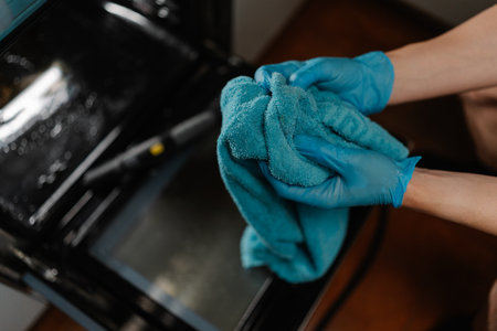 Top view of cleaning company worker wearing blue gloves squeezing out wet rag after cleaning inside of gas oven with steam cleaner on blurred background. Concept of housekeeping and cleaning.の写真素材