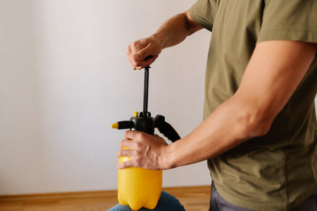 Janitor male pumping yellow water sprayer bottle while holding on blue sofa, preparing to clean or disinfect upholstery in home environment. Concept of domestic work, housekeeping and household.の写真素材
