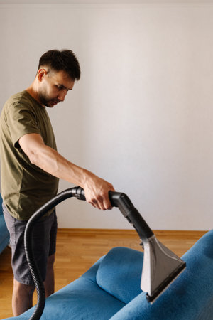 Vertical portrait of cleaning service worker using washing vacuum cleaner to remove dirt and stains from blue sofa in home or apartment, providing efficient and thorough upholstery cleaning, close-up.の写真素材