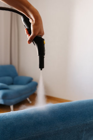 Homemaker using steam cleaning machine, removing tough stains from upholstered blue couch in modern living space, maintaining pristine furniture condition, close-up. Concept of cleaning and household.の写真素材