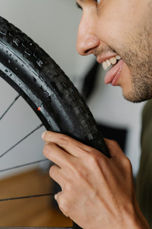 Close-up face of joyful man licking tire of mountain bike wheel while holding rag, showcasing humorous approach to bicycle maintenance, vertical portrait. Concept of bicycle transport service.の写真素材