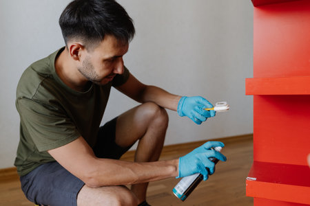 Professional cleaner male wearing blue gloves applying scotch remover spray and scrubbing red shelf with small brush, performing thorough housekeeping at home. Concept of hygiene and cleanliness.の写真素材