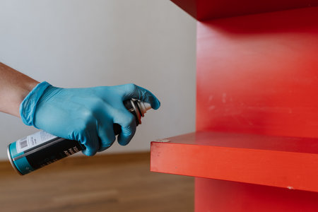 Close-up of cleaning company worker wearing blue gloves applying scotch remover spray to dried adhesive stain on red shelf, performing thorough housekeeping at home. Concept of hygiene and cleanlinessの写真素材