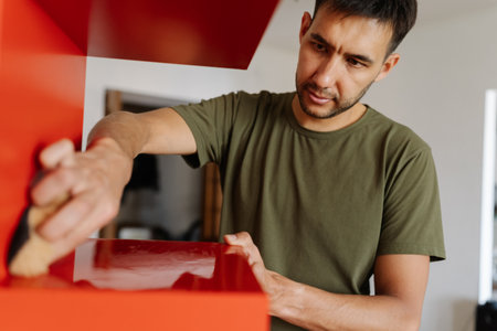 Portrait of young focused man cleaning wiping red shelf surface with sponge and detergent ensuring spotless and hygiene at home, close-up. Concept of cleanliness in domestic environment.の写真素材