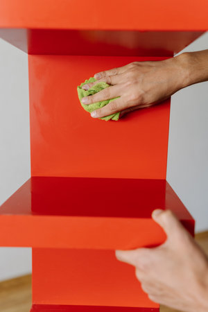 Cleaning service employee wiping red shelf with rag and detergent, ensuring spotless and dust-free surface in cleaning routine, close-up. Concept of domestic work, housekeeping and household.の写真素材