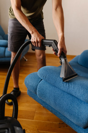 Close-up vertical cropped shot of cleaning service worker using washing vacuum cleaner to clean blue sofa in modern living room. Concept of hygiene and cleanliness in domestic environment.の写真素材