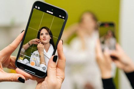 Professional beautician applying makeup while female hands recording process, creating social media beauty tutorial with smartphone in modern beauty clinic. Concept of anti-age rejuvenation skincare.の写真素材