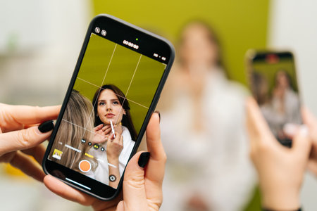 Female hands holding smartphone recording video of beautician applying makeup on model face, creating content for social media or online tutorial in cosmetology clinic. Concept of beauty treatment.の写真素材