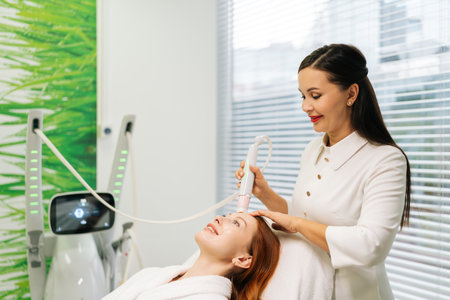 Beautician using modern device performing mesotherapy treatment on forehead of young woman lying on treatment table in modern beauty salon with light interior. Concept of healthcare technology.の写真素材