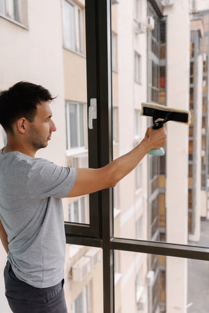 Vertical shot of young man spraying cleaning solution onto large window and wiping clean with squeegee, ensuring spotless and streak-free finish in building. Concept of cleanliness and housekeeping.の写真素材