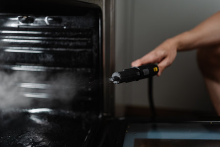 Closeup of professional cleaner worker using steam cleaner removing stubborn grease and grime from modern gas oven interior, delivering thorough kitchen appliance sanitization.の写真素材