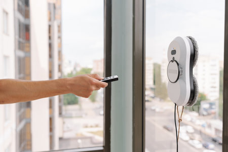 Close-up cropped shot of housekeeper using remote controller to operate robotic window cleaner, ensuring efficient and safe cleaning of high-rise building windows with cityscape view.の写真素材