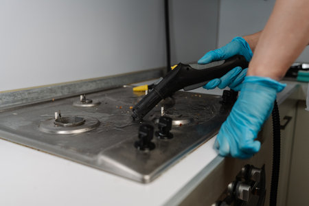 Cropped shot of housewife wearing blue gloves using steam cleaner to remove burned-on food and grease from dirty gas stove, ensuring hygiene and cleanliness in kitchen setting, close-up.の写真素材