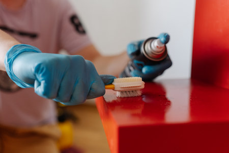 Cropped shot of housewife male wearing blue protective gloves removing adhesive stains, scrubbing red wooden shelf with small brush during detailed cleaning process at home, close-up.の写真素材