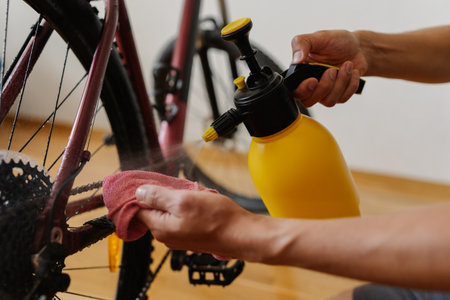Detail cropped shot of unrecognizable man using yellow sprayer and cloth preparing to clean chain of mountain bike, emphasizing importance of bicycle maintenance after trail riding at home.の写真素材