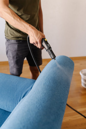 Vertical cropped shot of cleaning service employee sanitizing blue sofa with high-temperature steam cleaner in contemporary living space, removing dirt and bacteria. Concept of hygiene and cleanlinessの写真素材